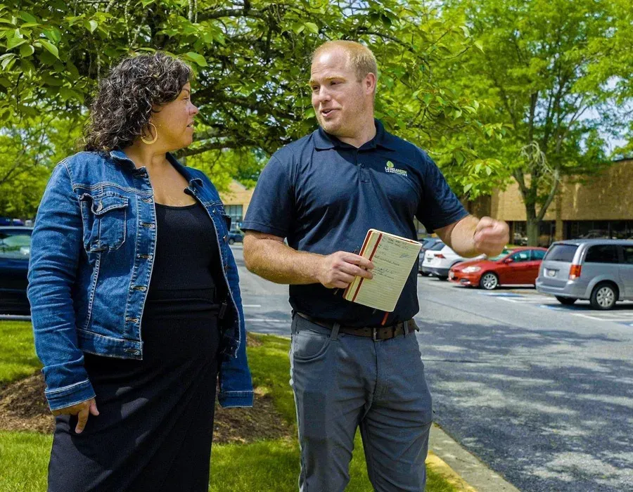Two people talking on a tree-lined sidewalk, one holding a clipboard near parked cars.