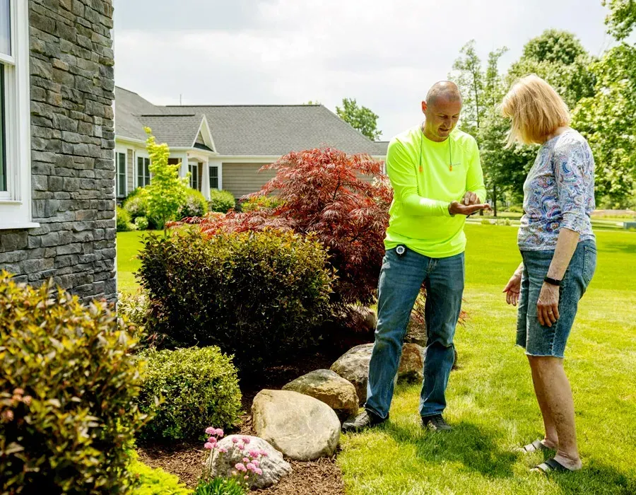 Two people talking in a garden beside shrubs and a stone house, with one holding a device