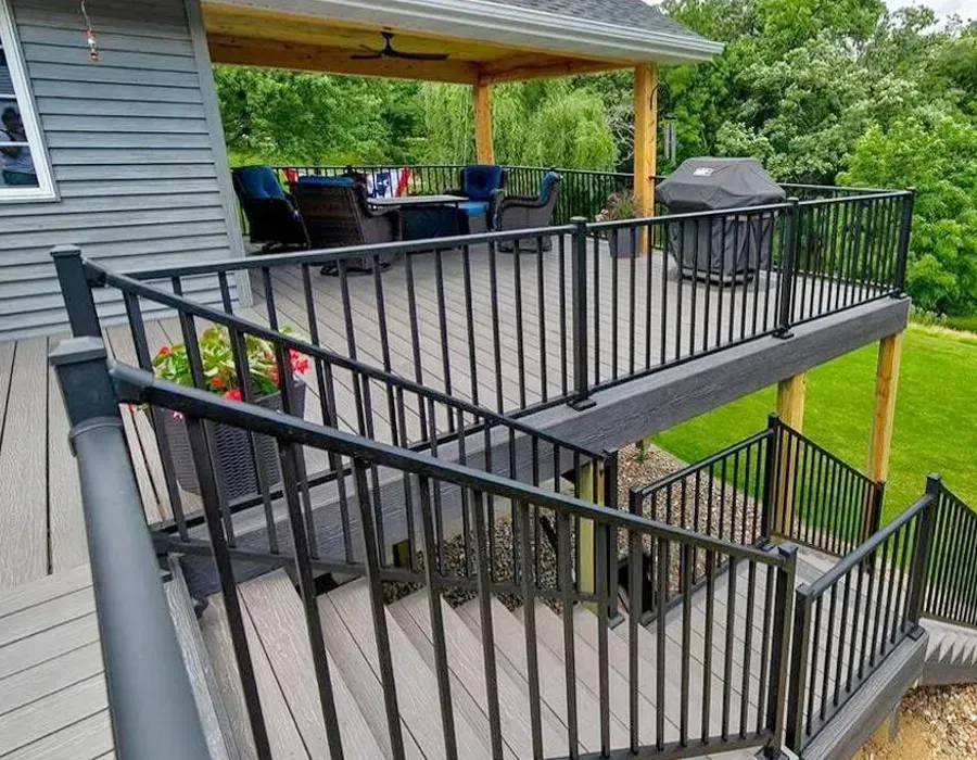 Two-tier wooden deck with black railings and stairs beside a house, with a covered patio and grill.