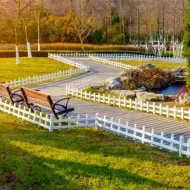 Winding park path with white fence, wooden bench, pond, and autumn trees in bright sunlight