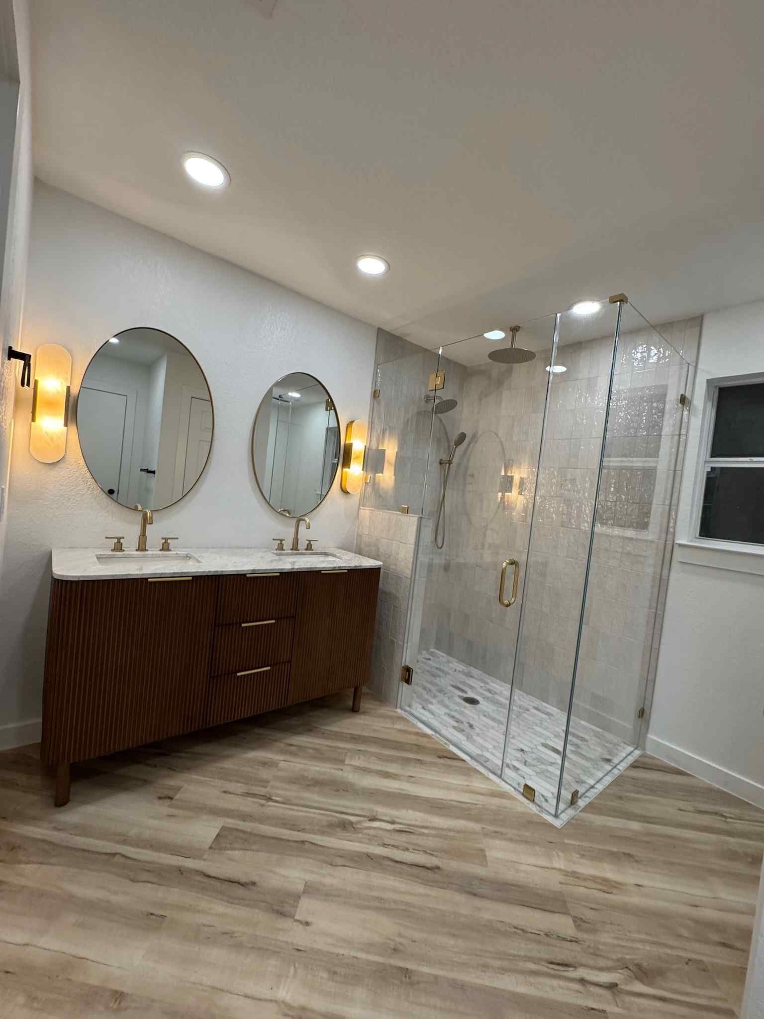 Modern bathroom with double vanity, oval mirrors, and glass shower enclosure on light wood flooring.