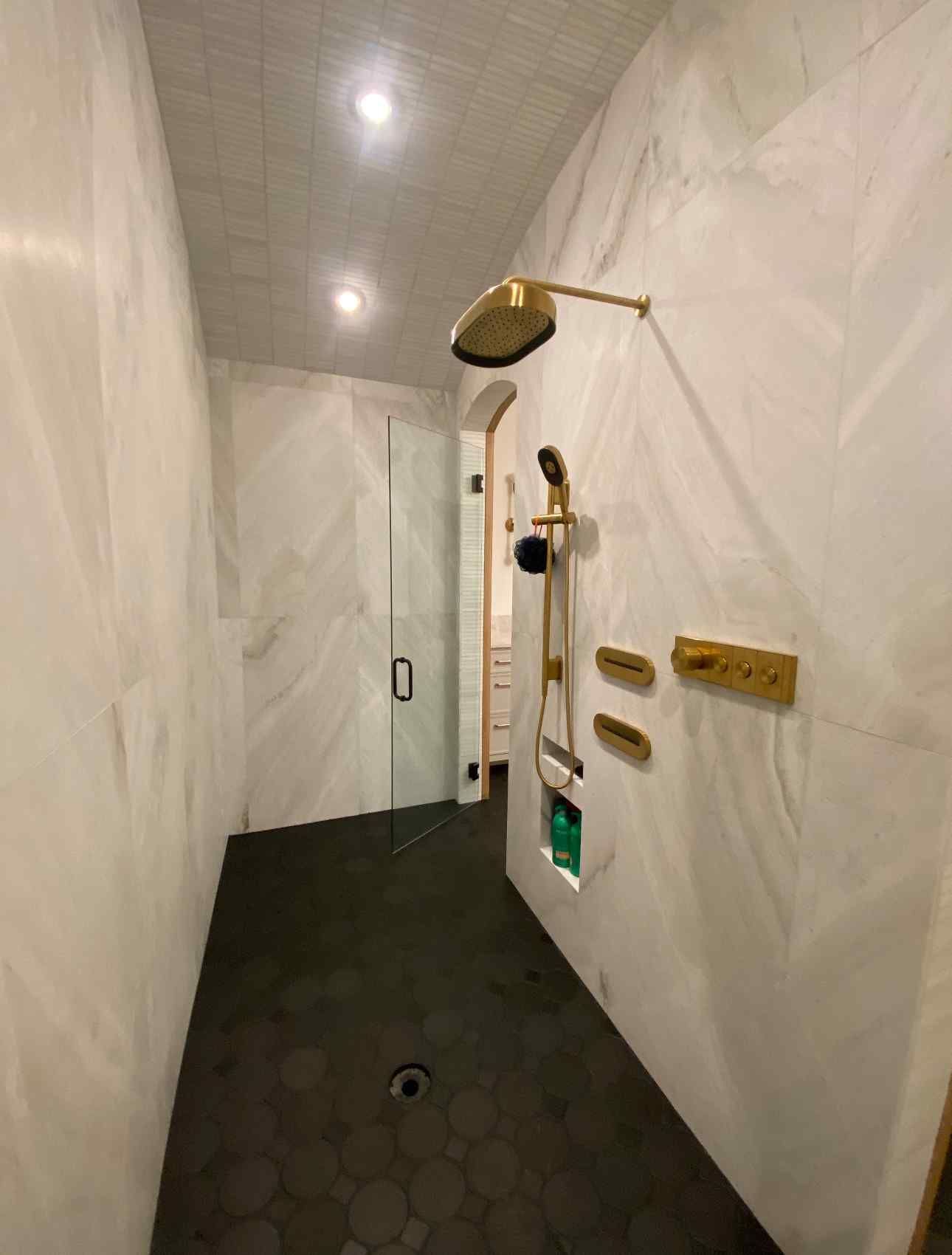 Modern marble shower with dark hex tile floor, brass fixtures, and glass door in a narrow bathroom.