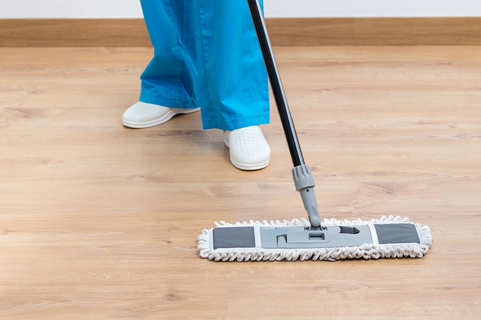 Person mopping a light wood floor with a gray mop while wearing blue pants and white shoes