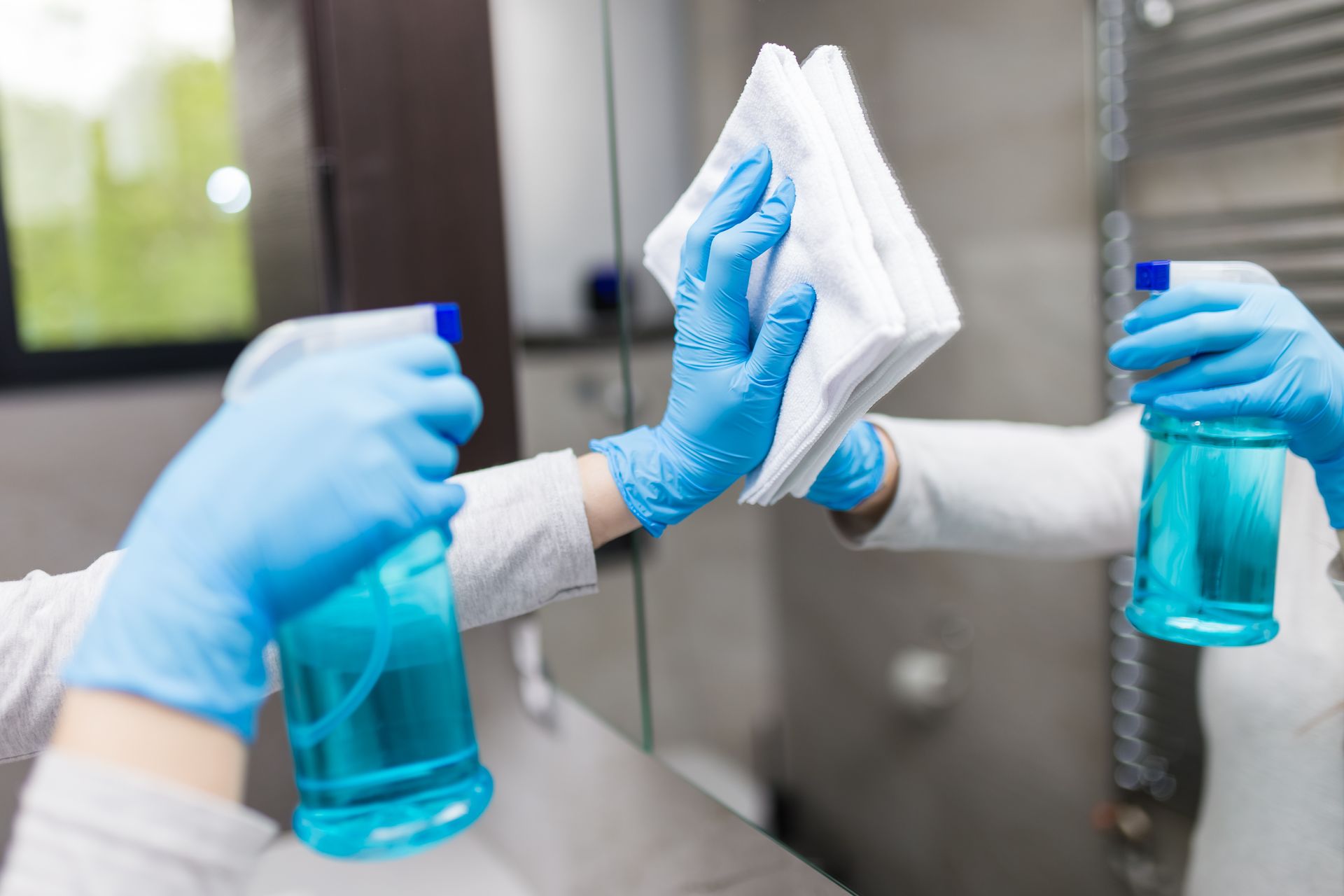 Gloved hands wiping a mirror with a white cloth while holding blue spray bottles in a bright restroom
