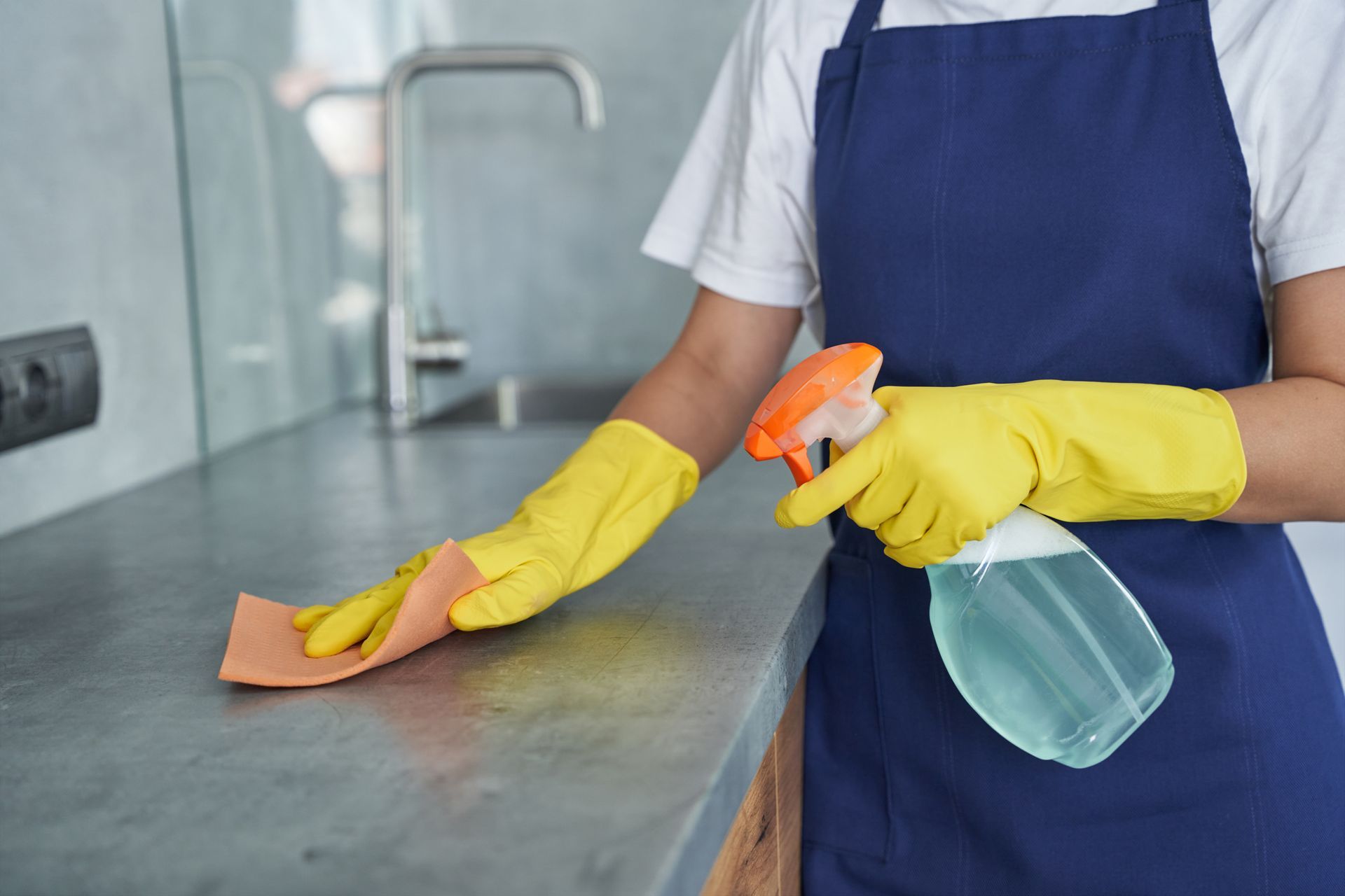 Person in blue apron wearing yellow gloves sprays and wipes a kitchen counter with a cloth.