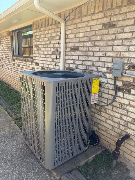 A gray, box-shaped air conditioning unit sits on a concrete path outside a light-colored brick building.