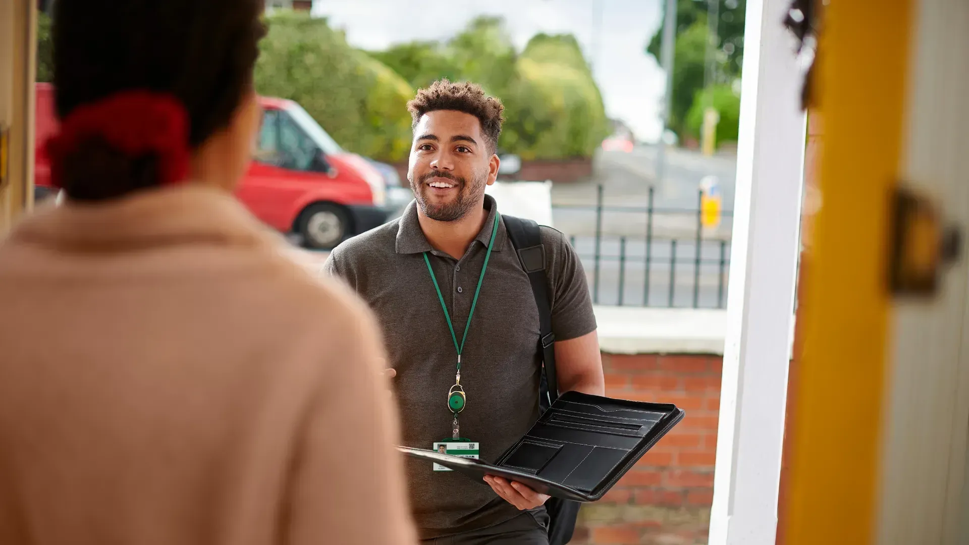 Person at doorway speaking with another. Man holds tablet, wearing lanyard. Red van in background.