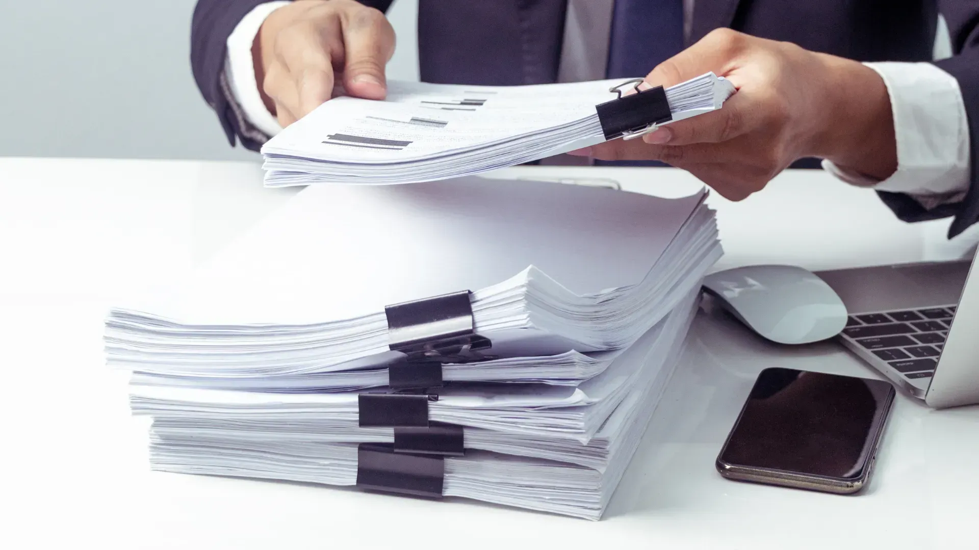 Person in suit arranging a stack of paper documents held together with binder clips on a desk with a phone and mouse.