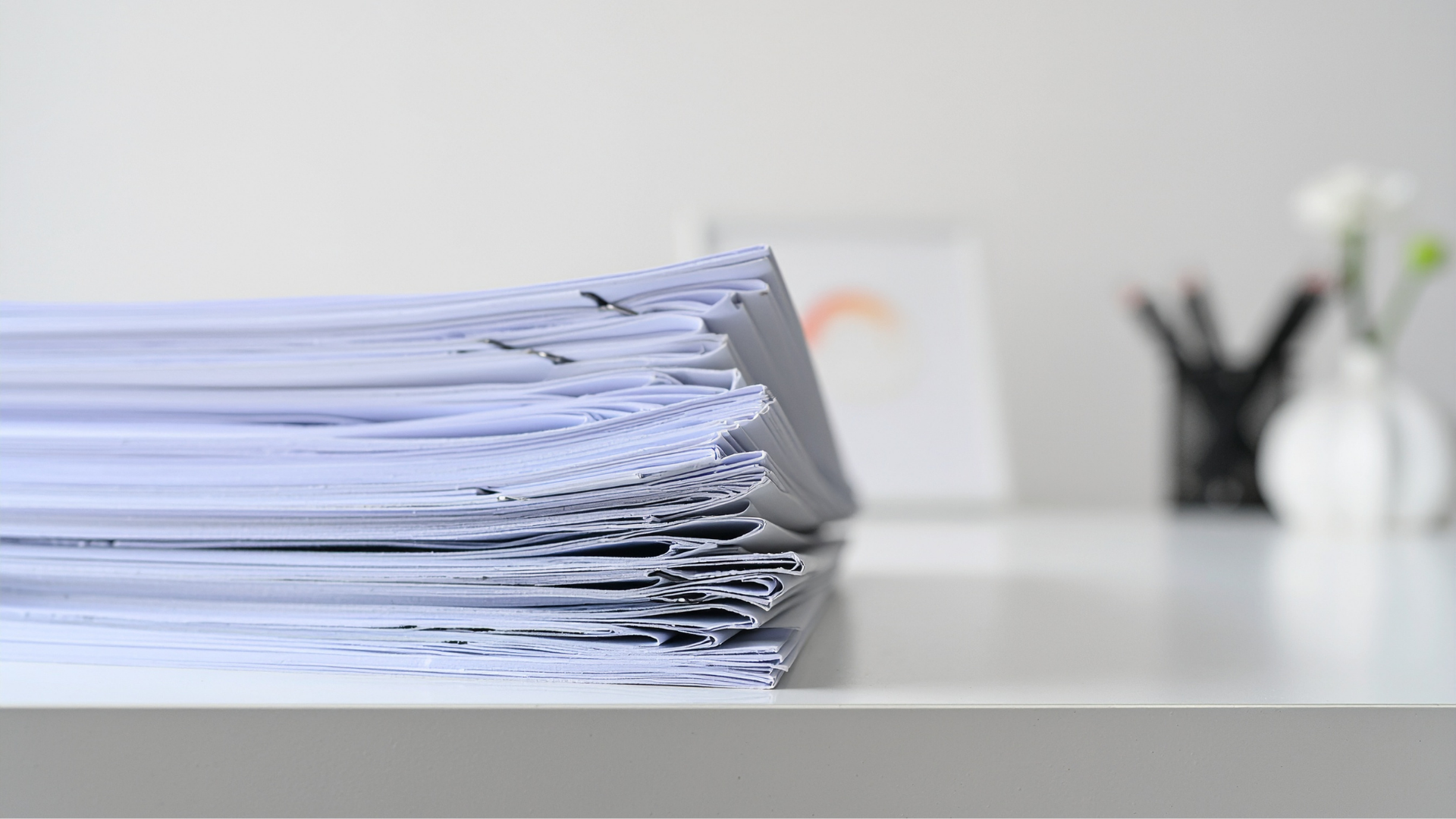 Pile of white paper on a white desk with blurred office supplies and a vase of flowers in the background.