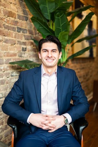 Man in a blue suit, sitting, smiling, hands clasped. Brick wall and plant in background.