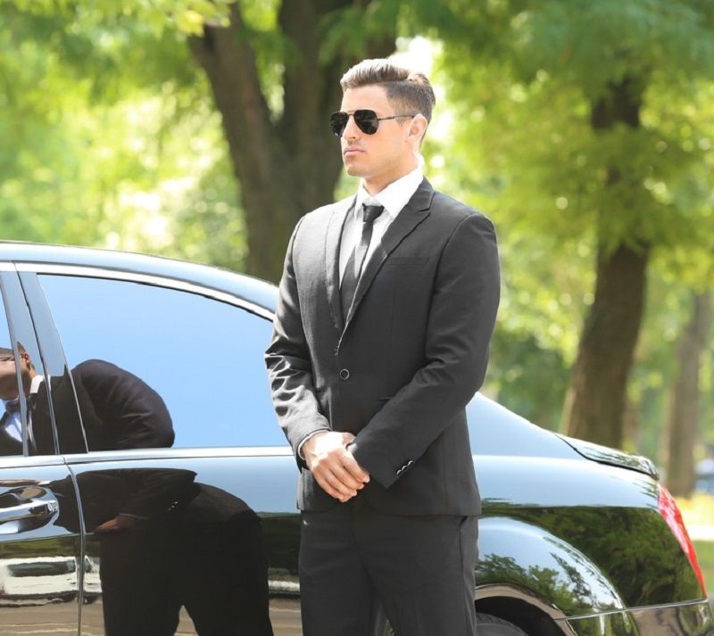 Man in black suit and sunglasses stands near a black car, in a park-like setting.