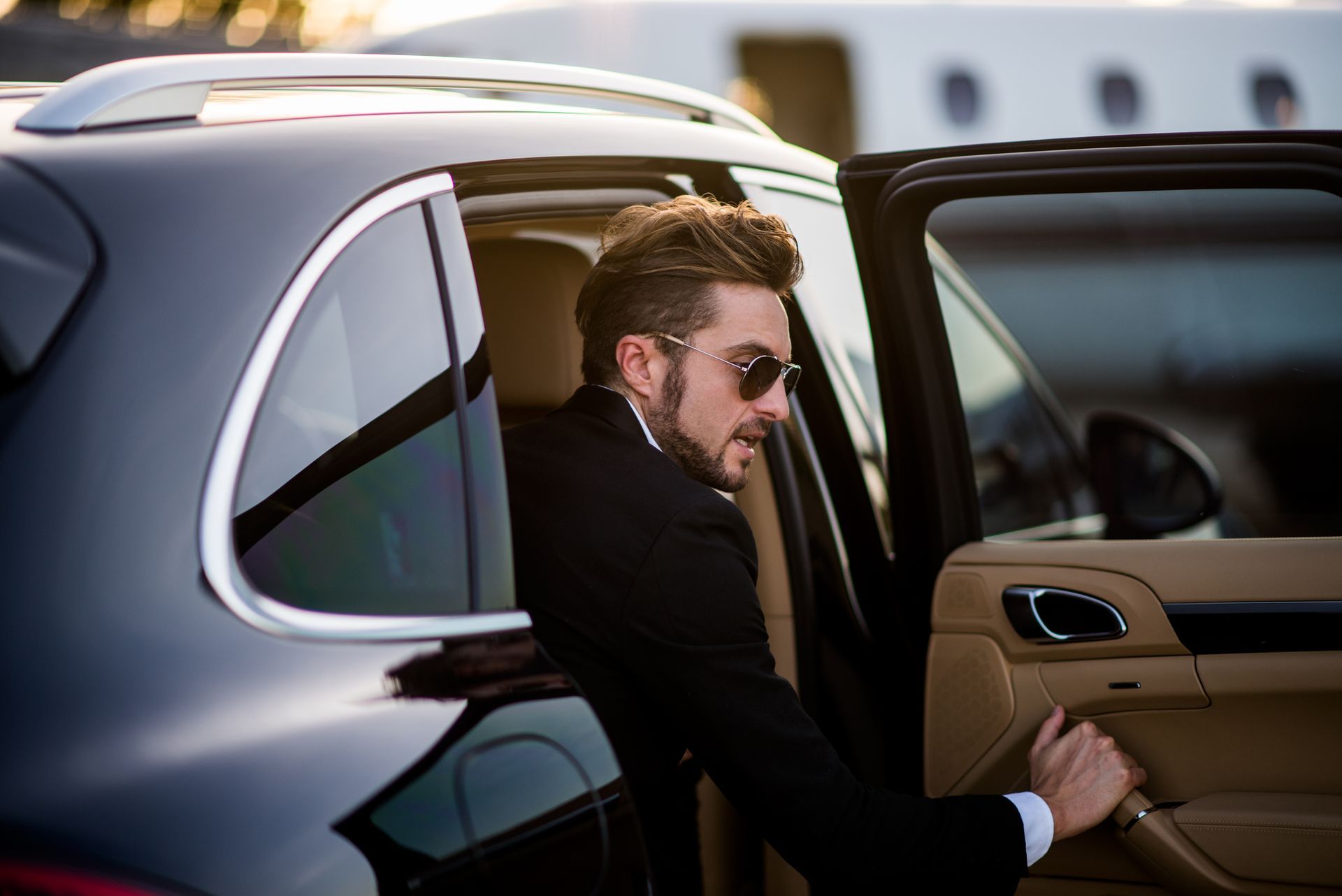 Man in a suit exits a black car near a private jet. He wears sunglasses.