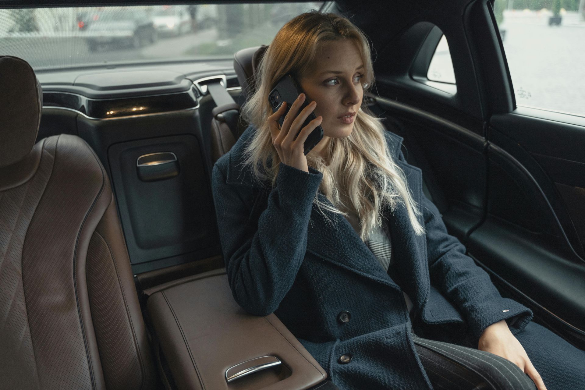 Woman in a car talking on the phone, looking concerned. Dark blue coat, brown leather seats.