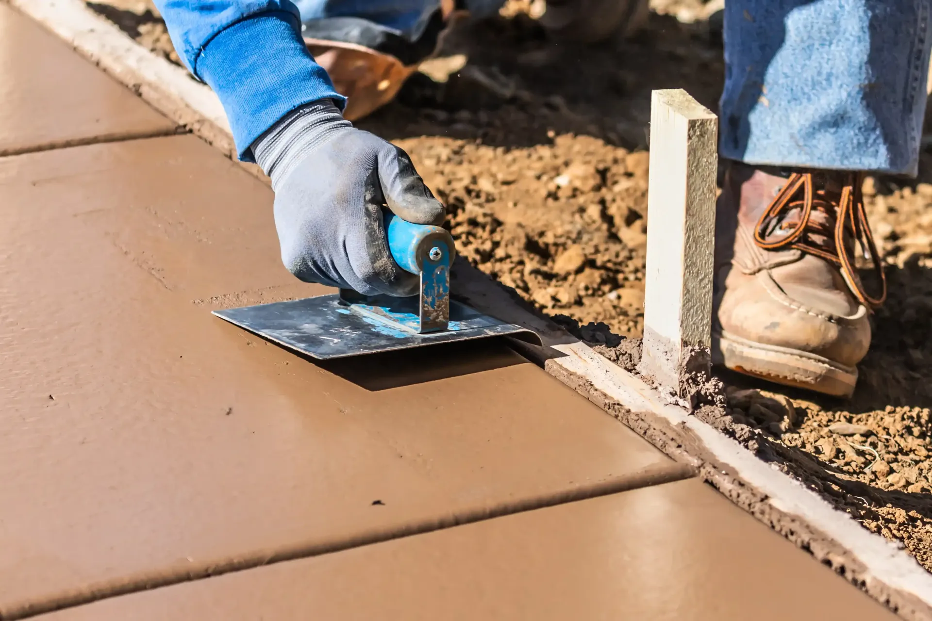 A worker smoothing wet concrete with a trowel, creating a sidewalk; outdoor construction.