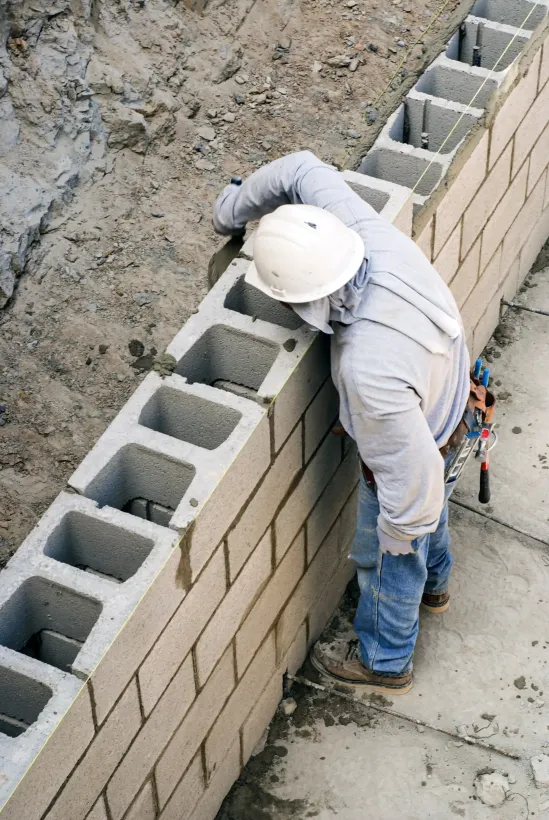 Construction worker wearing a white hard hat building a cinder block wall outdoors.