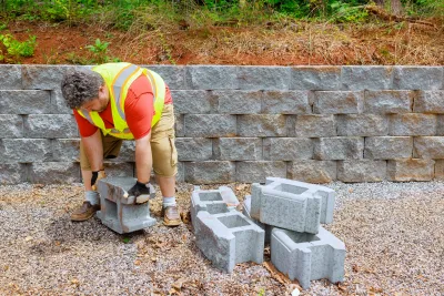 Man in safety vest stacking concrete blocks to build a retaining wall outdoors.