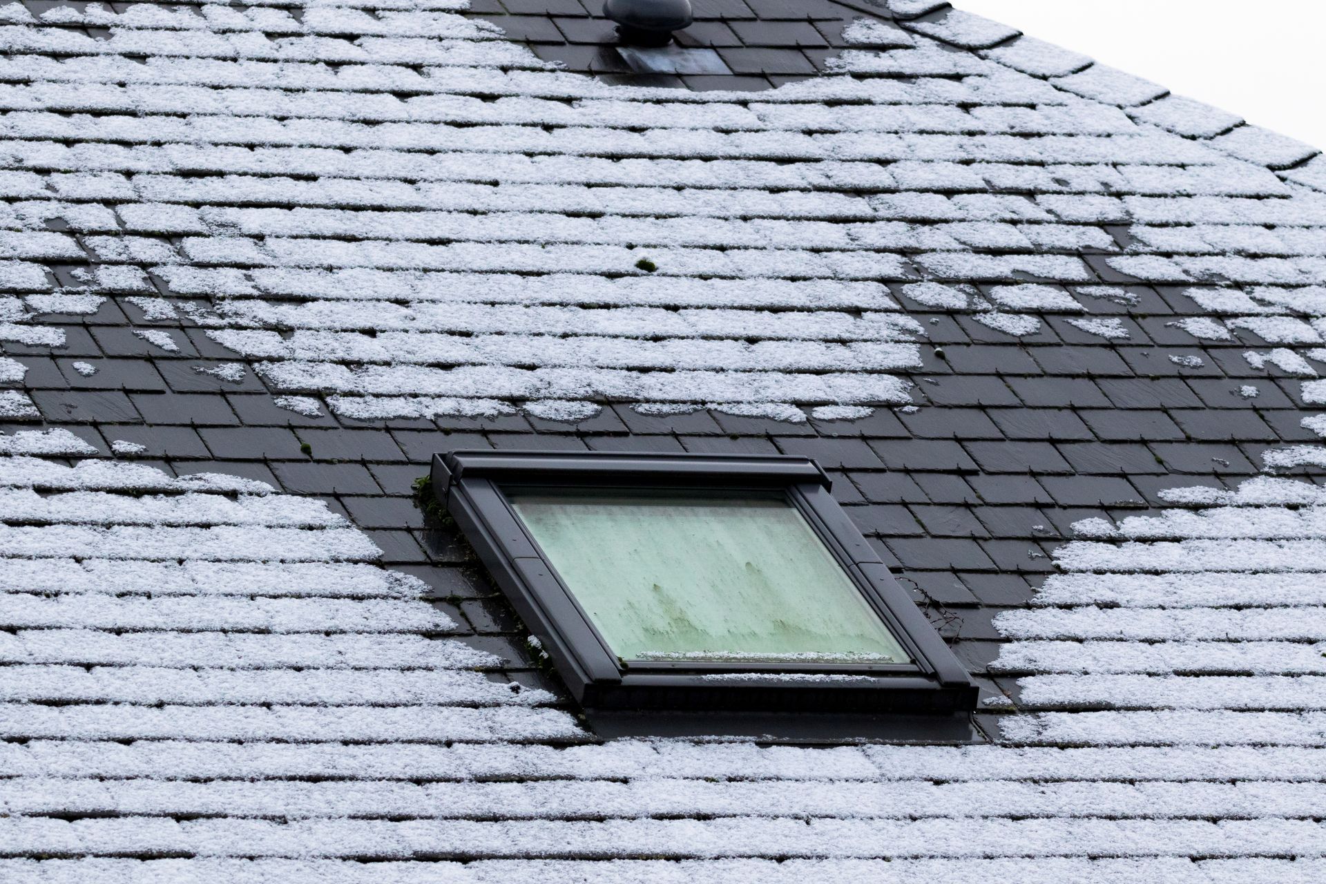 Snow-covered slate roof with a skylight; melting snow revealing the dark tiles.