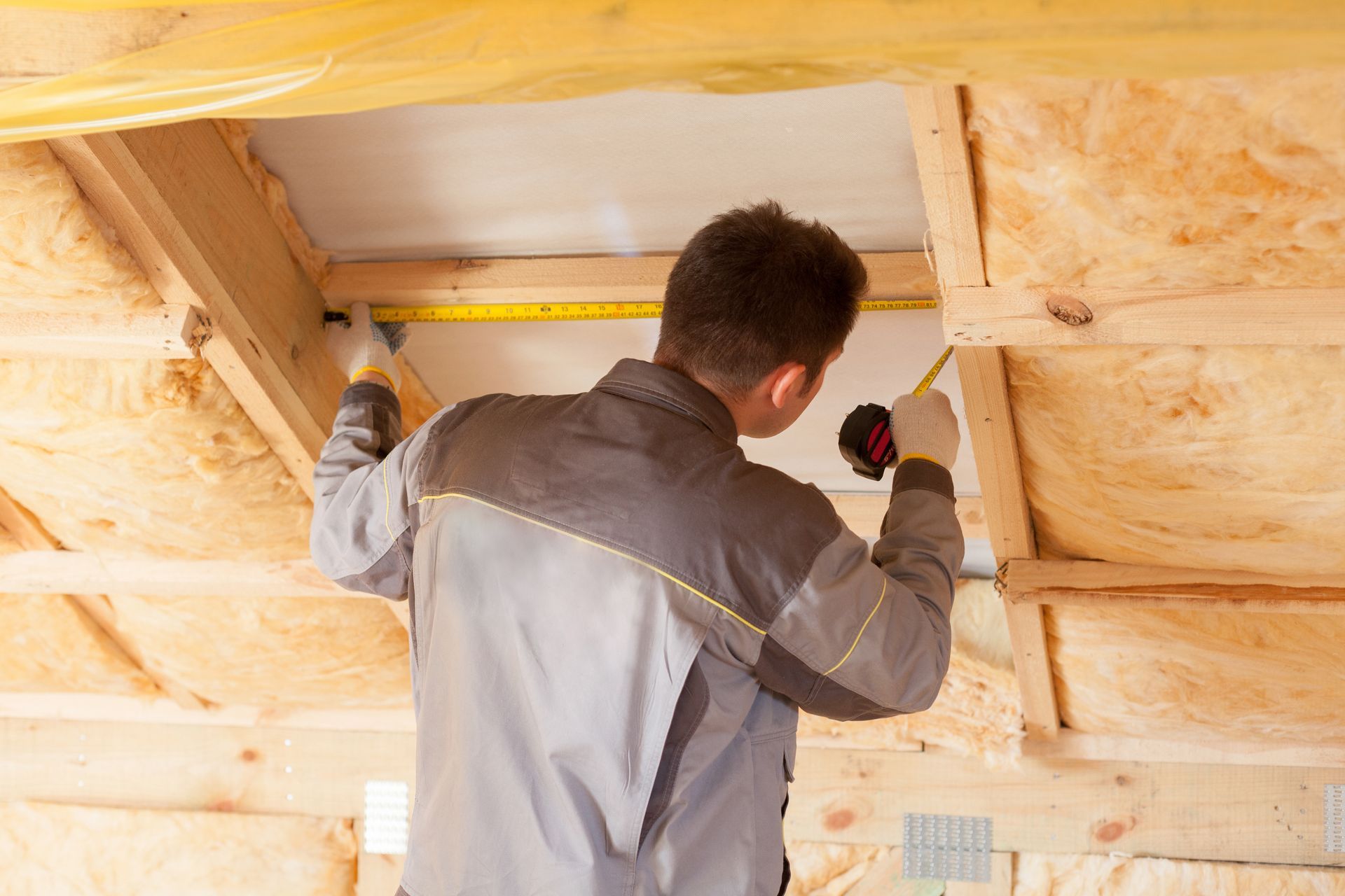 Construction worker measures with tape inside a wood-framed structure with insulation.