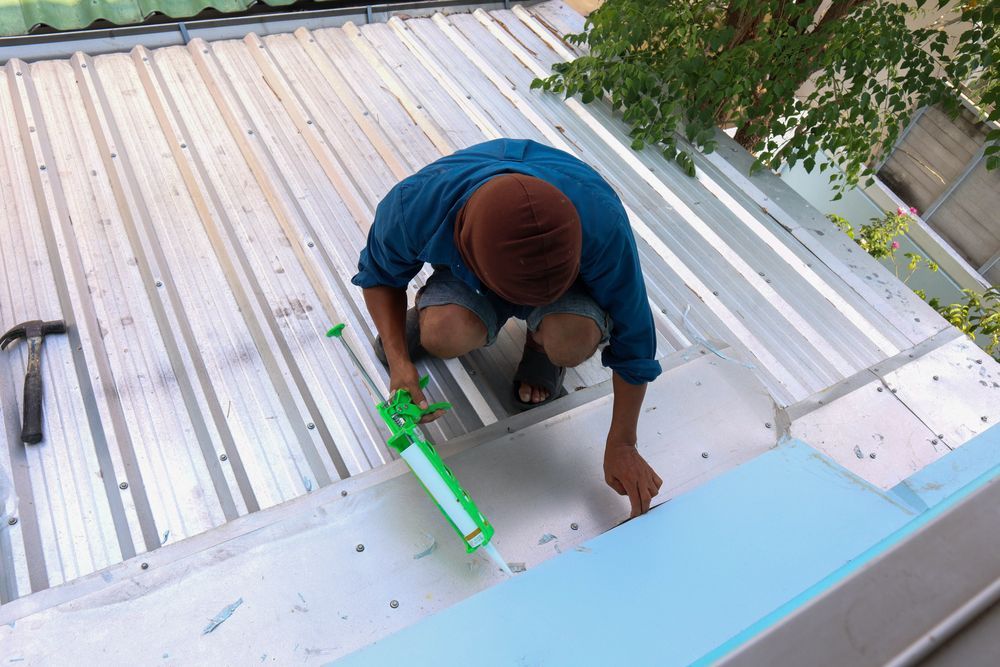 A man is kneeling on a roof holding a green sealant.
