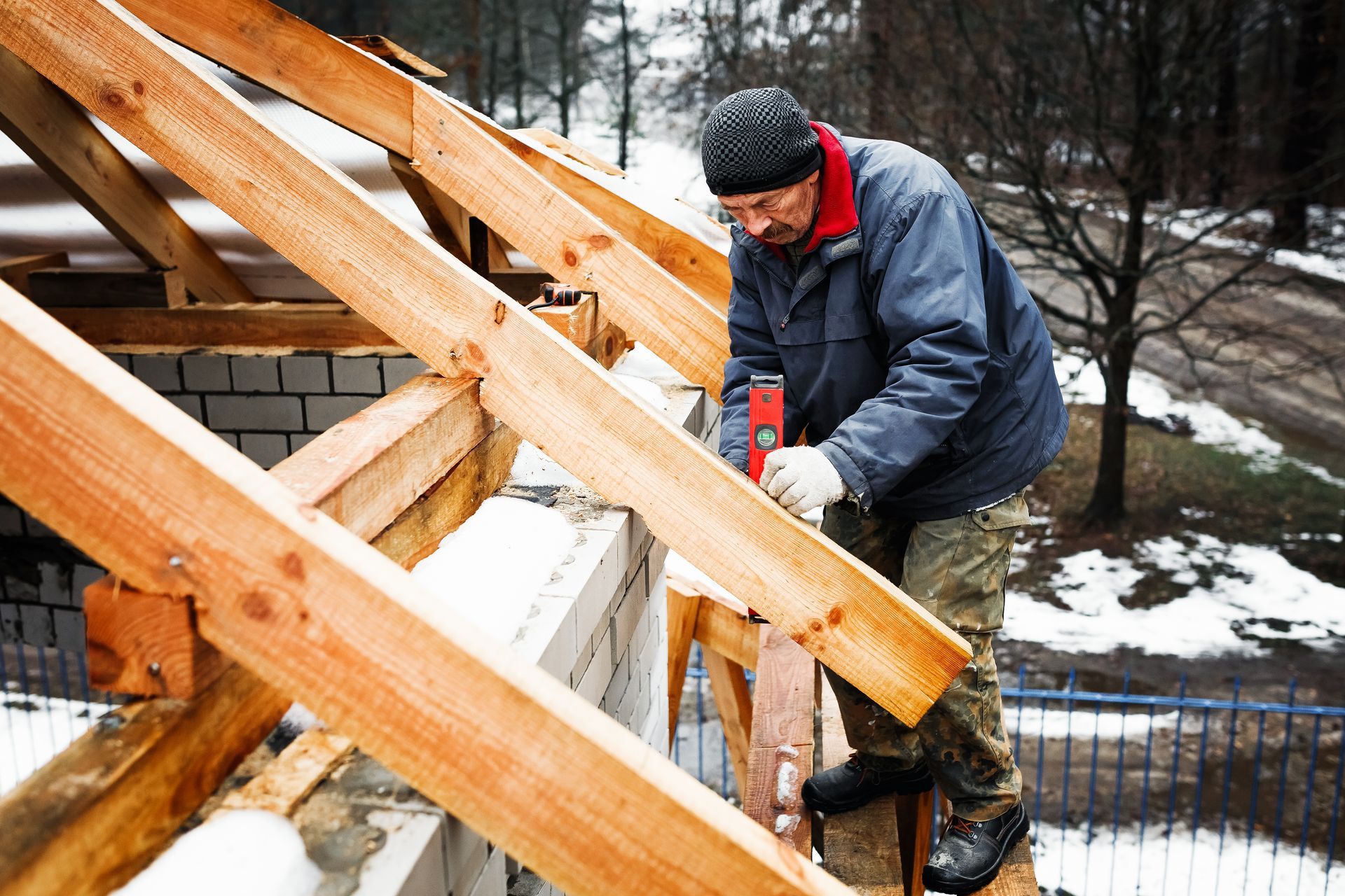 Carpenter in winter clothes using a level on roof beams. Snow is visible in the background.