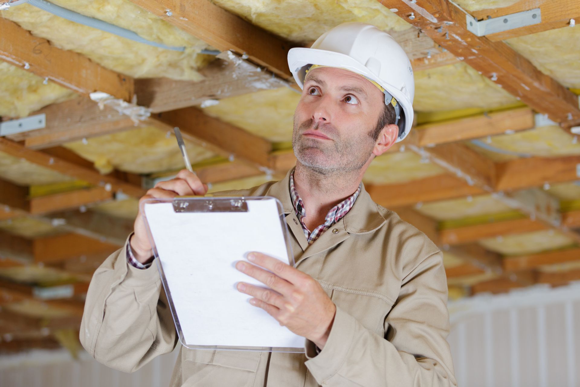 Man in a hard hat inspecting a ceiling, taking notes on a clipboard.