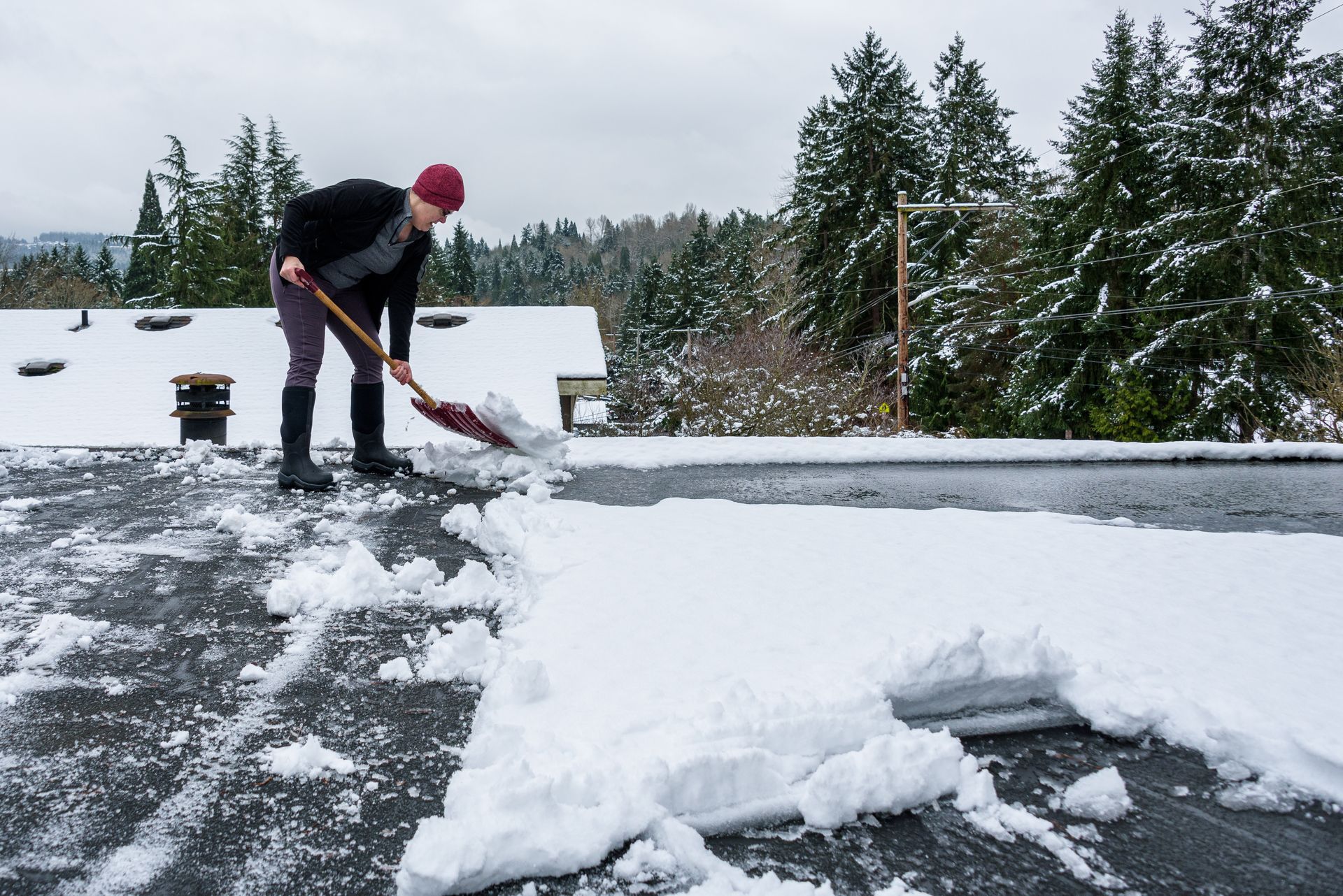 Person shoveling snow from a roof, wearing a black jacket and red hat. Overcast day, trees in the background.