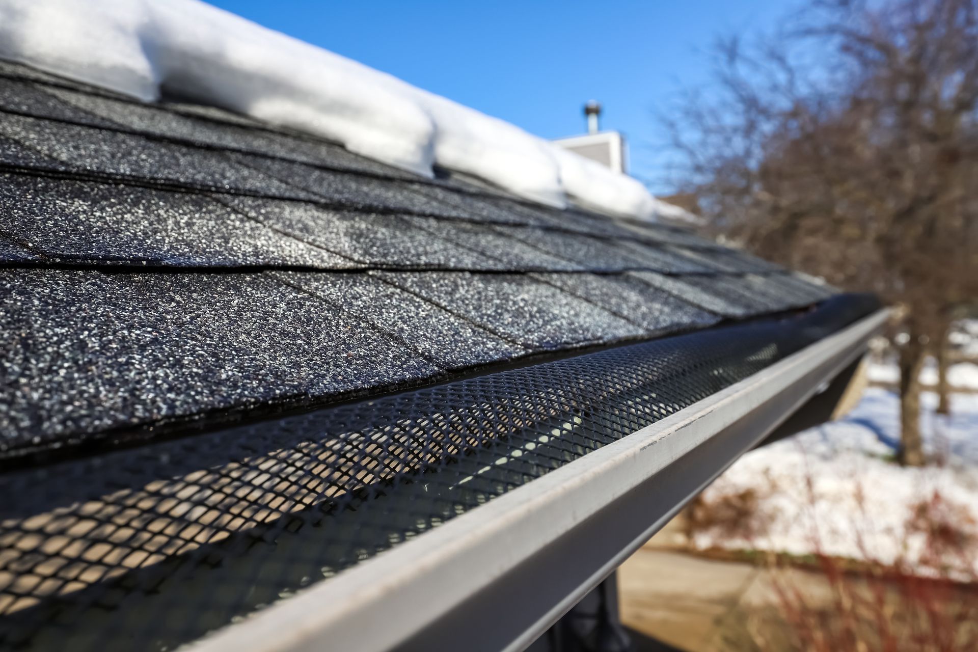 Close-up of a rooftop with snow, a gutter with mesh guard, and a blue sky in the background.