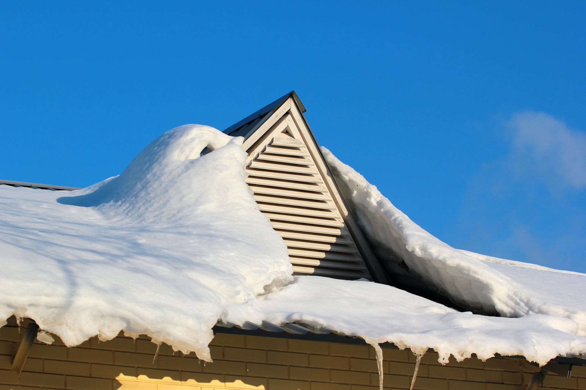 Snow-covered roof with a triangular vent against a clear blue sky.