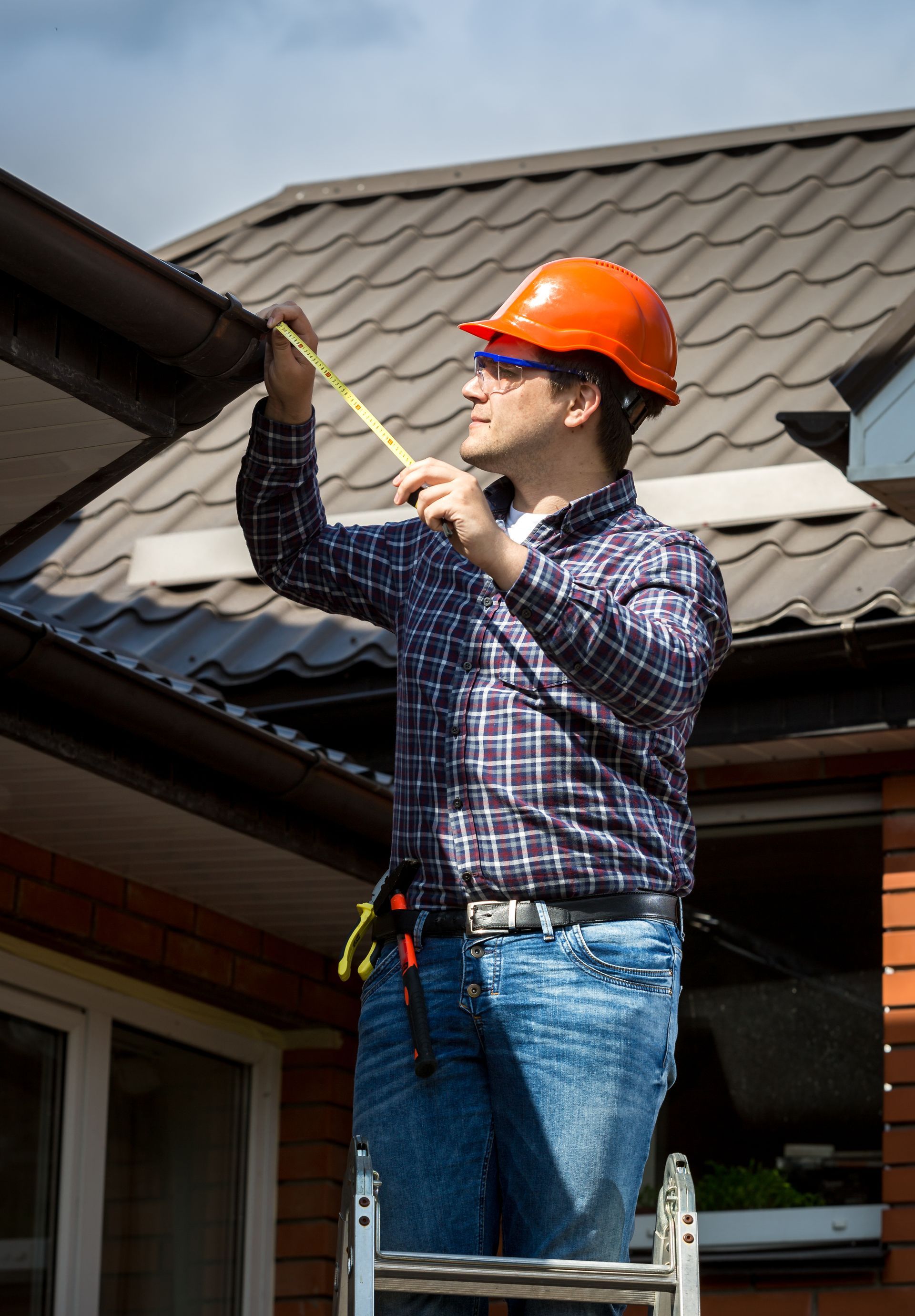 A man is standing on a ladder measuring a gutter with a tape measure.