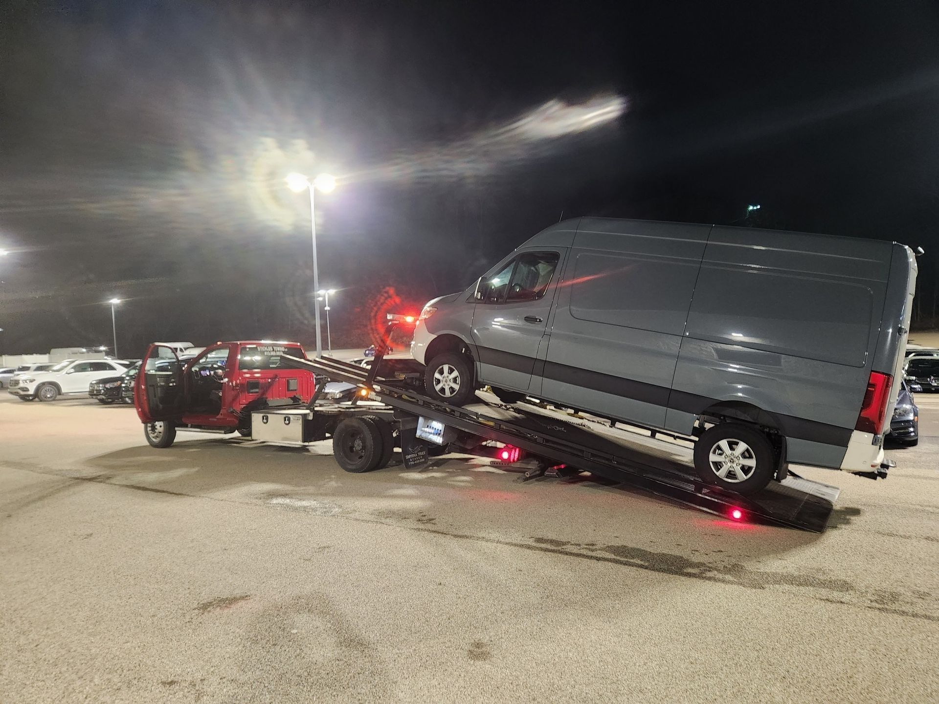 A gray van is being towed by a tow truck in a parking lot at night.