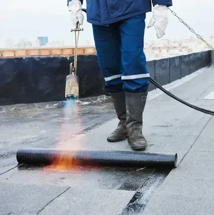 Une personne portant une veste bleue et des bottes brûle du matériau de toiture sur un toit plat.