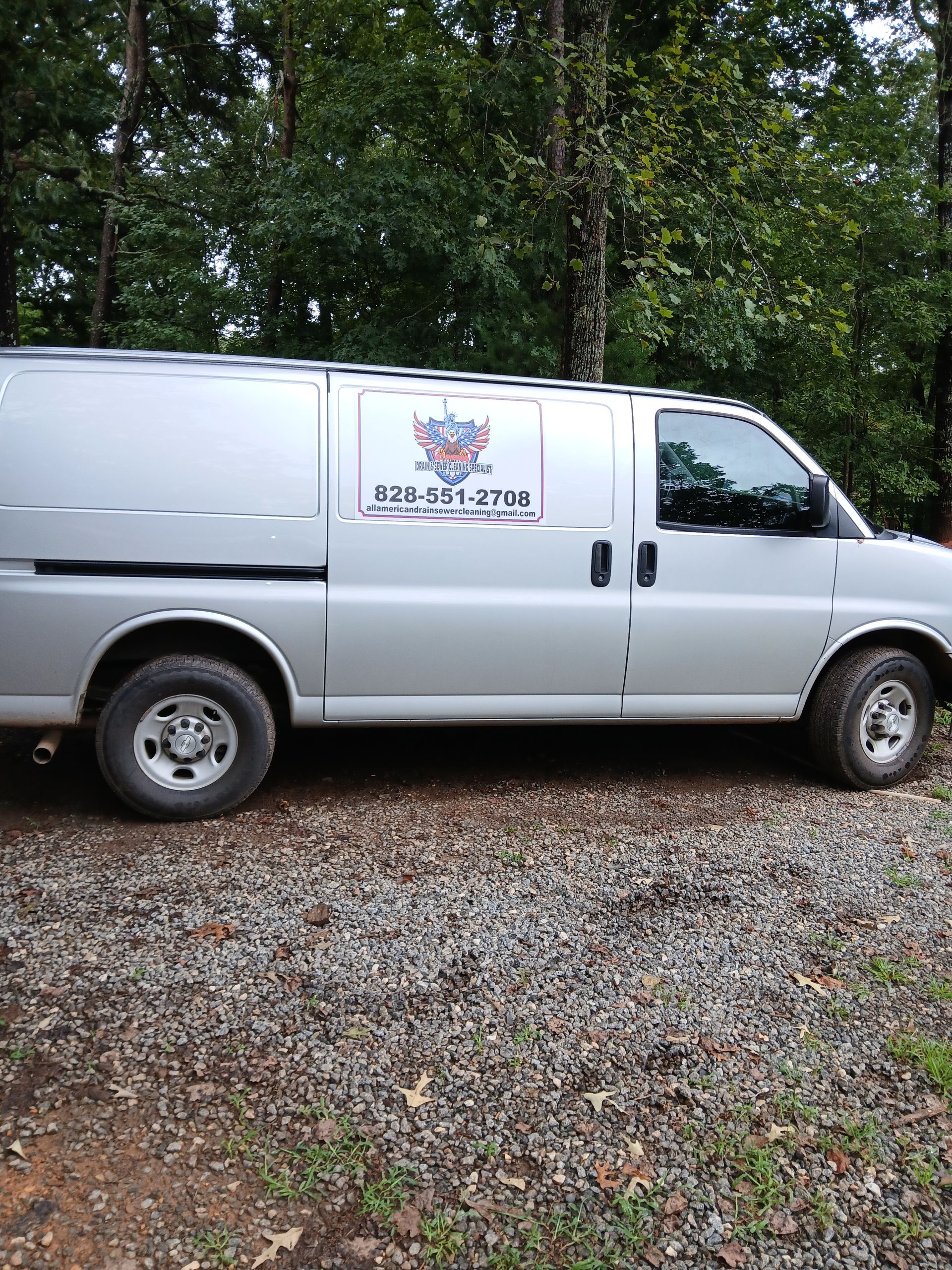 Silver service van parked on gravel, with logo and phone number on the side. Outdoors, trees in the background.