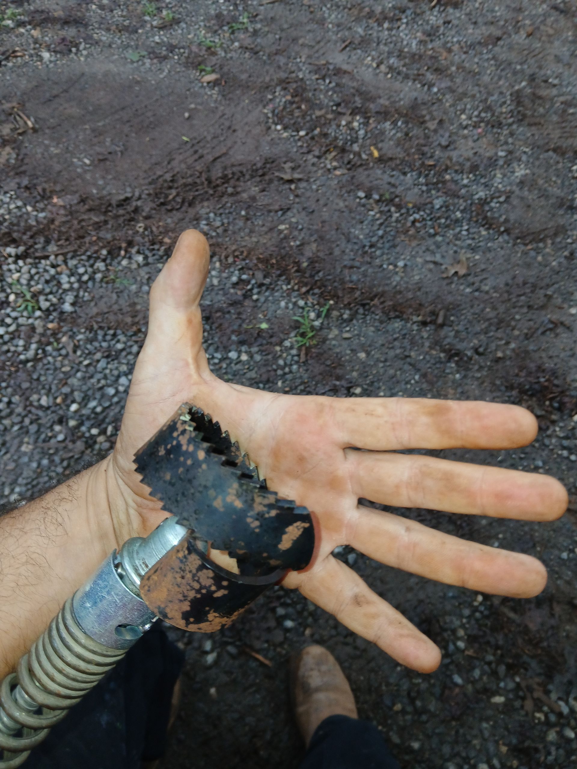 Hand holding a dirty, toothed blade attached to a hose, outdoors on muddy ground.