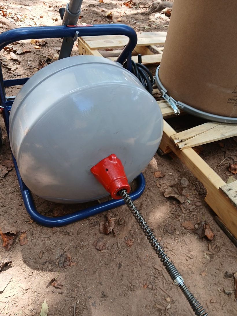 Plumbing snake machine with a cable extending. Grey drum, blue frame, red cable end. Outdoor setting.