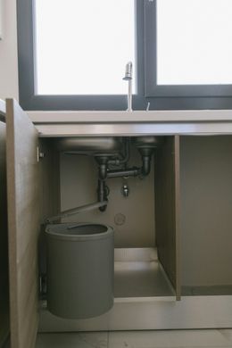 Under-sink view: stainless steel sinks, plumbing, waste bin, and cabinet door.