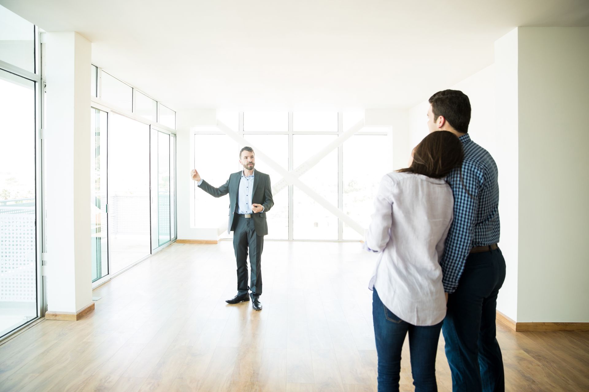 Three people in a bright empty room listening to a man in a suit point toward the windows