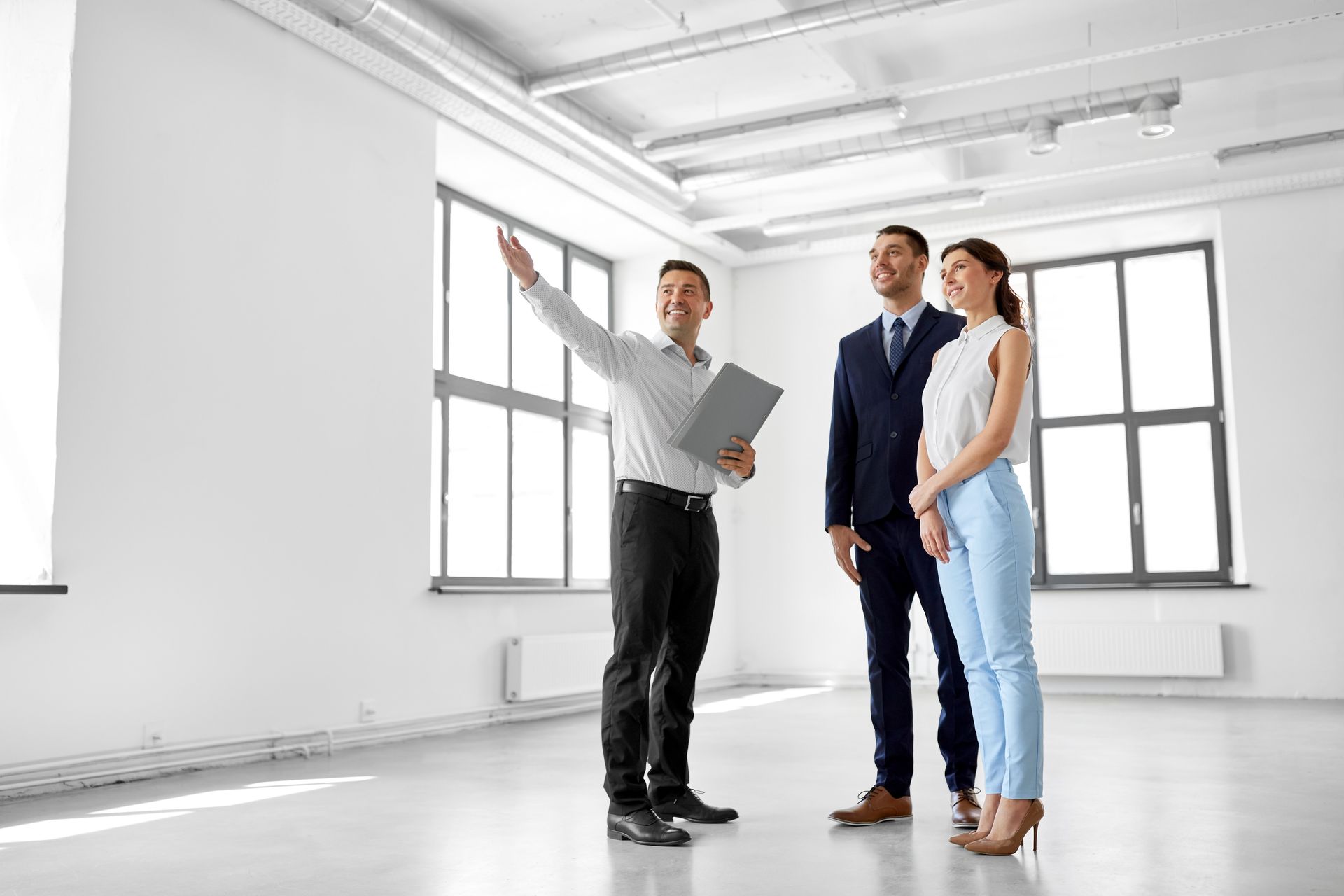 Three people in business attire touring a bright empty room with large windows