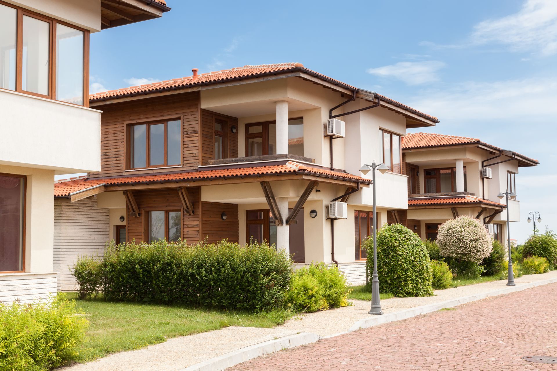 Row of modern beige townhouses with red-tiled roofs and green shrubs along a paved street