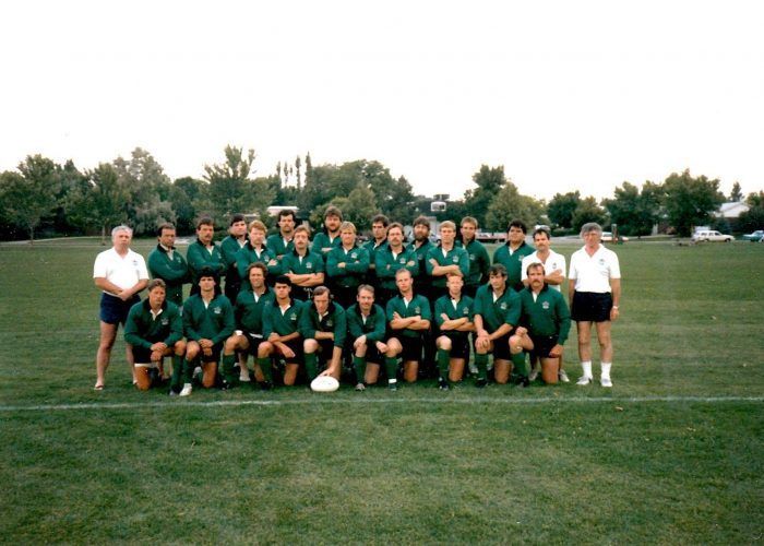 A group of rugby players in dark green uniforms pose for a team photo on a grassy field with two coaches on the ends.