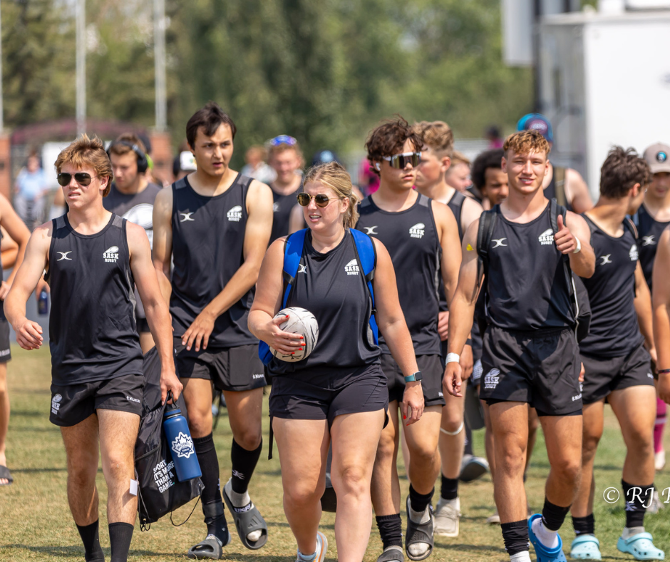 A group of athletes in black team uniforms walking together on a grassy field, some carrying sports equipment.