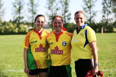 Three people smile while wearing sports jerseys on a grassy field.