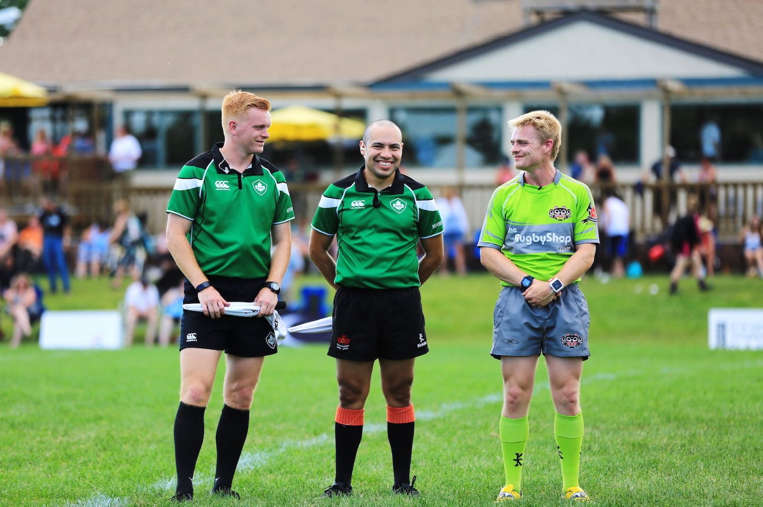Three match officials in athletic uniforms stand side-by-side on a grassy field, smiling during an outdoor sporting event.