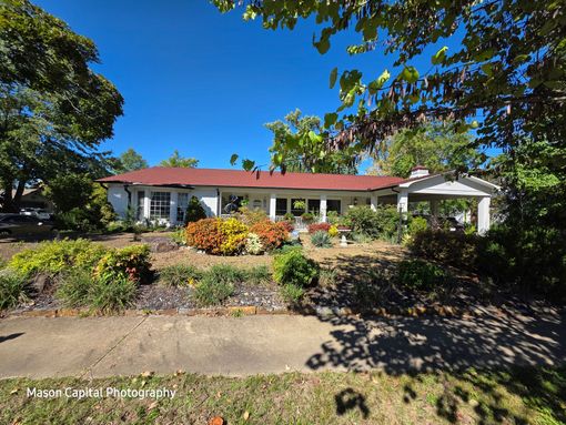 White, single-story house with a red roof, surrounded by a garden and trees under a clear blue sky.