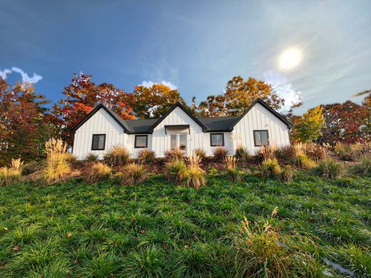 White farmhouse with black trim, surrounded by colorful fall foliage, set on a grassy hill under a sunny sky.