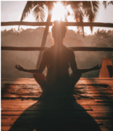 A person sitting cross legged facing a sunrise with arms  perched either side meditating.