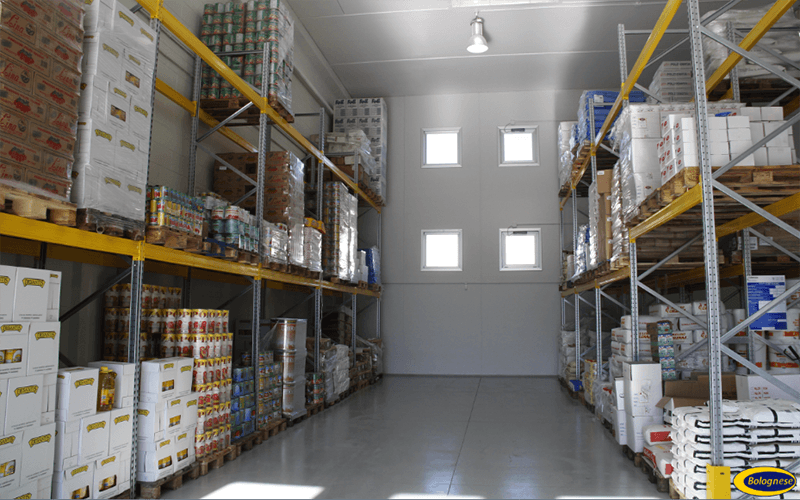 Interno di un magazzino con scaffali pieni di merci imballate e pallet. Pareti bianche, pavimento grigio, luce intensa dall'alto.