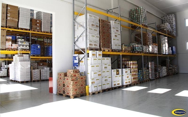 Interno di un magazzino con scaffali gialli pieni di scatole, pallet e merci, sotto luci intense.