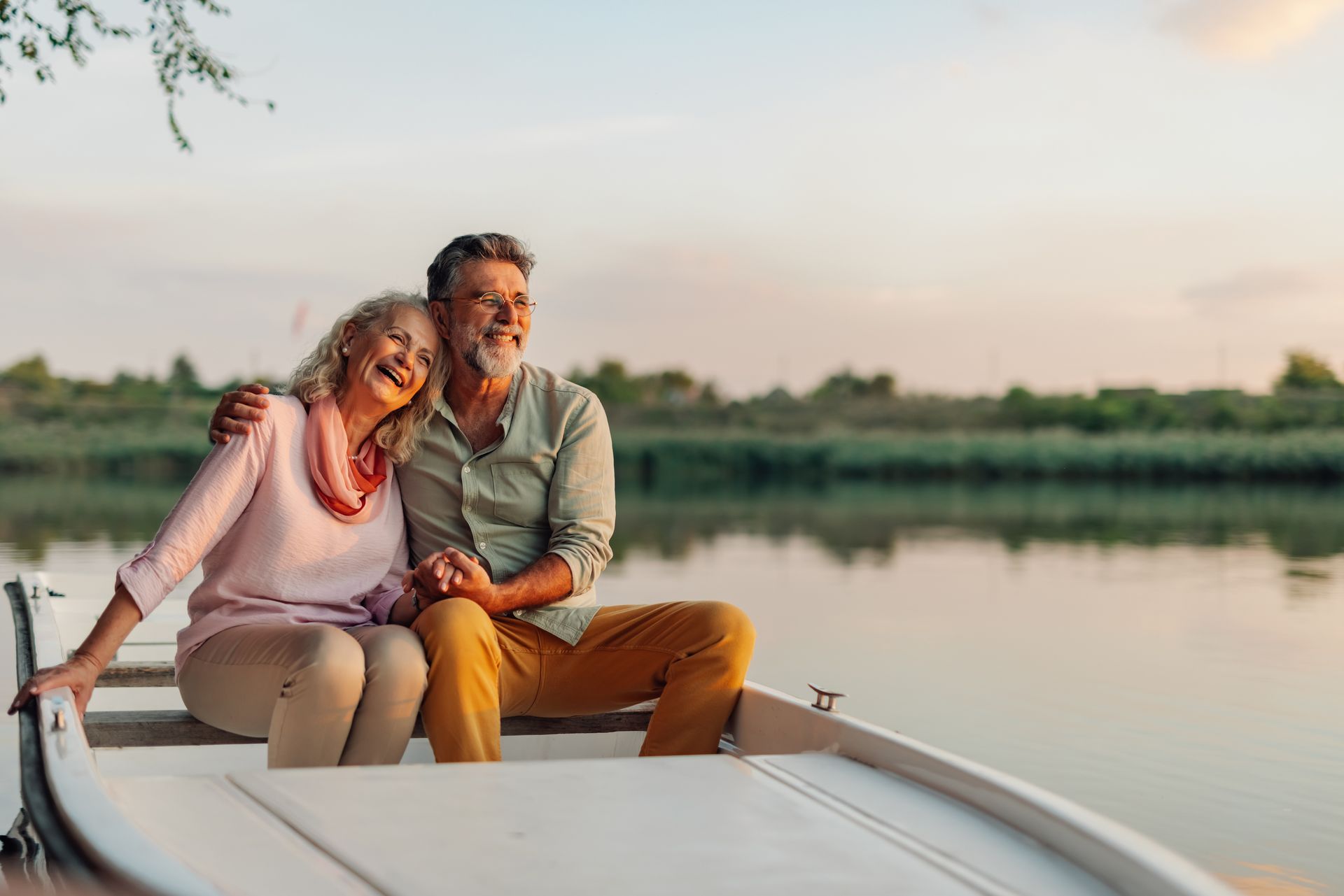 Couple in a boat on a lake at sunset, smiling, holding hands.