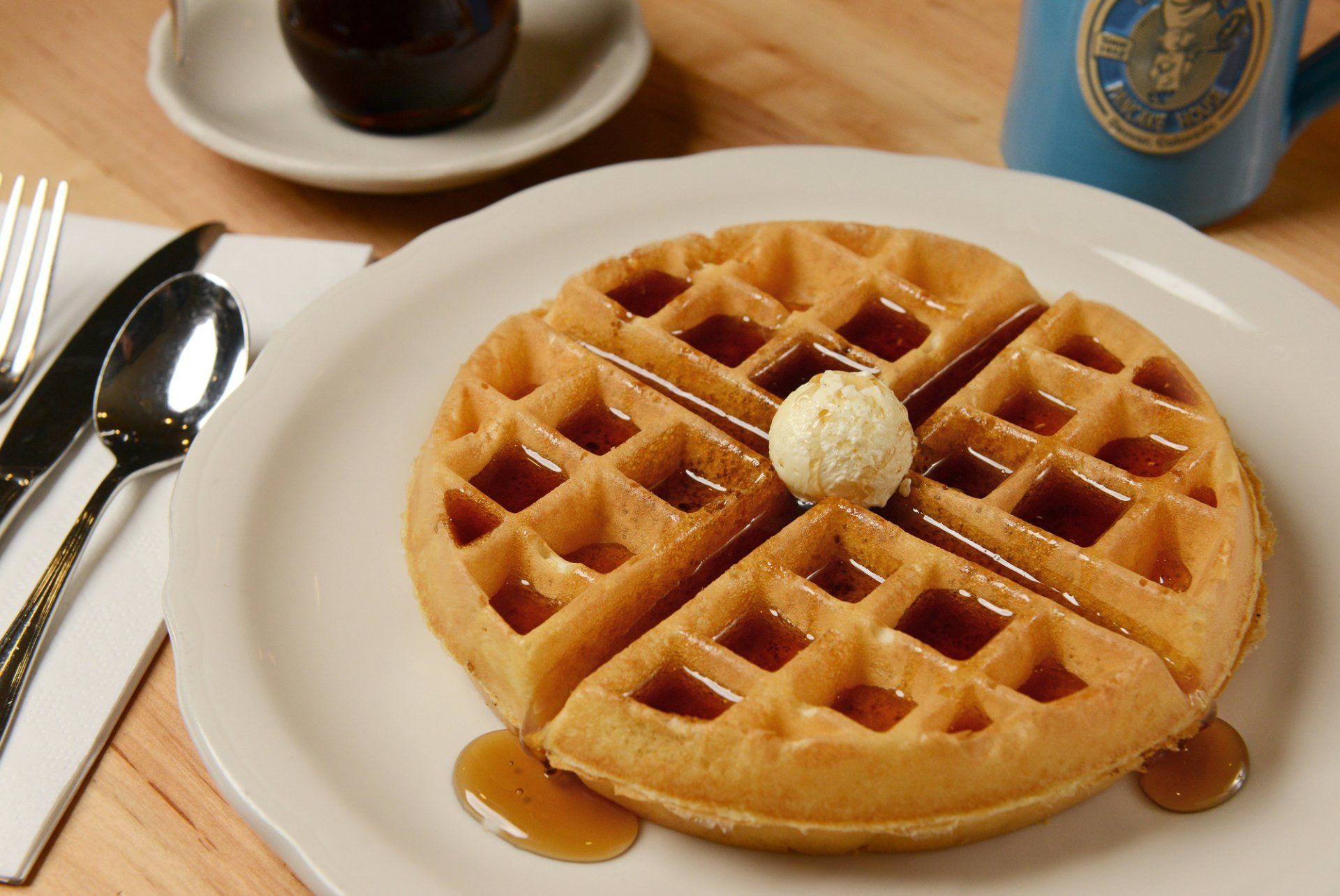 Waffle with butter and syrup on a white plate, with silverware and a mug in the background.