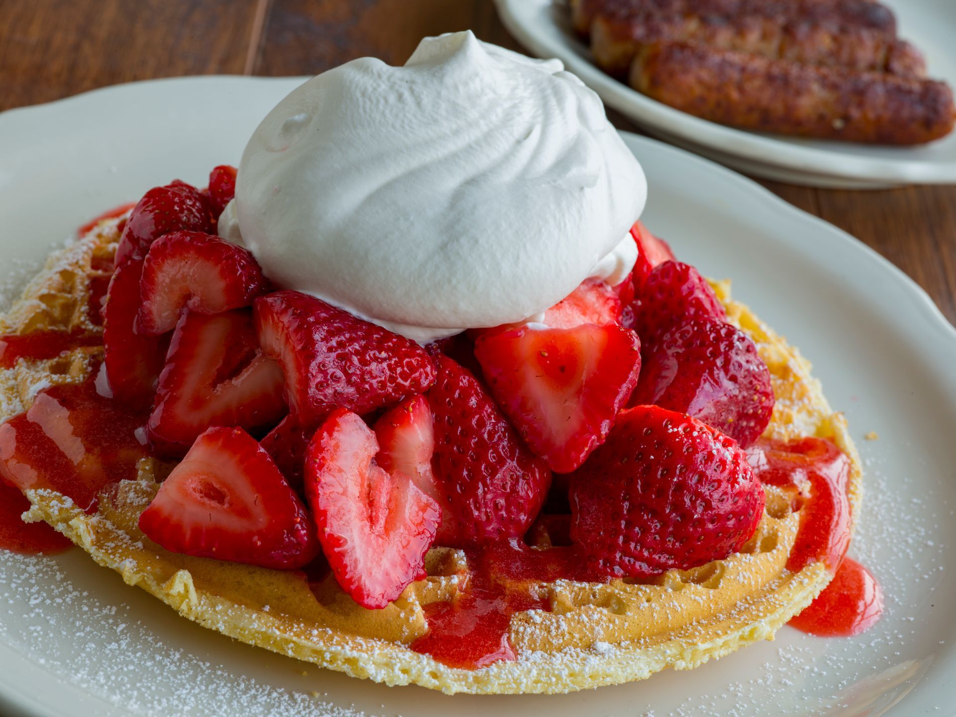 Waffle topped with sliced strawberries, whipped cream, and sauce on a white plate, with sausage in the background.